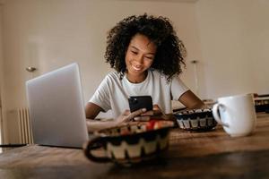 Black young woman smiling and using mobile phone with laptop and breakfast photo