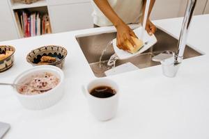 Joven negra lavando platos mientras usa el portátil en la cocina foto