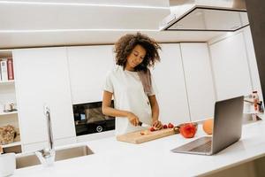 Black young woman making salad while using laptop in kitchen photo