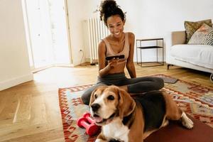 Black young woman using cellphone while sitting with her dog on mat photo