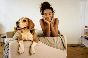 Black young woman laughing while resting with her dog on her sofa photo