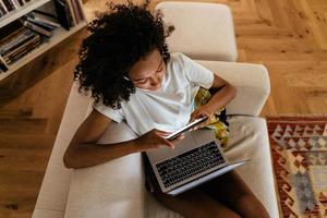Black young woman using mobile phone and laptop while resting on sofa photo