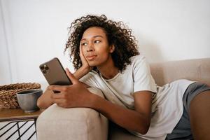 Black young woman using mobile phone while resting on sofa looking to the side photo