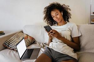 Joven negra en auriculares con teléfono móvil mientras descansa en el sofá