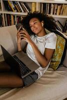Joven negra con teléfono móvil y portátil mientras descansa en el sofá