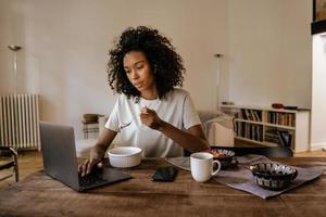 Black young woman using laptop and drinking coffee while sitting at table