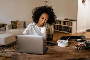 Black young woman using laptop and drinking coffee while sitting at table