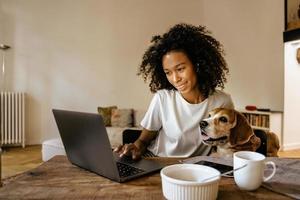 Black woman using laptop and hugging her dog while having breakfast photo