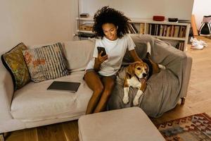 Joven negra con teléfono celular y acariciando a su perro en el sofá foto