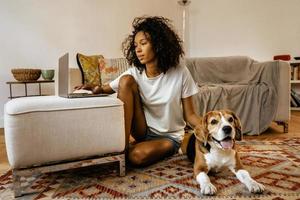 Mujer negra con laptop y acariciando a su perro mientras está sentada en el piso foto