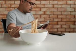 Asian male worker reads an appointment book while eating instant noodles in white bowl with chopsticks on table in brick wall background office during a lunchtime break, a hastily unhealthy lifestyle. photo