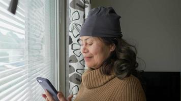 Aziatische oude vrouw die naar het raam loopt, door het raam naar buiten kijkt en vervolgens op de telefoon typt en het uitzicht naar buiten ziet. winter seizoen. quarantaine concept video