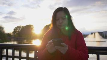 close-up aziatische vrouw met een rode jas die bij de rivier in het park staat en op haar telefoon typt om een praatje te maken met een vriend, mooie dag met rivier en zonsondergang op de achtergrond, fijne avond video