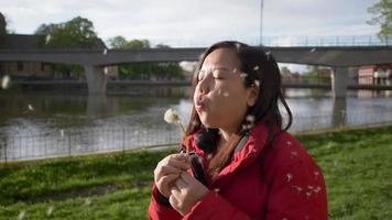 mujer asiática soplando polen en el parque el fin de semana, teniendo un momento de relajación y un momento feliz. tomando aire fresco con hermosa vista, pasto verde y río video