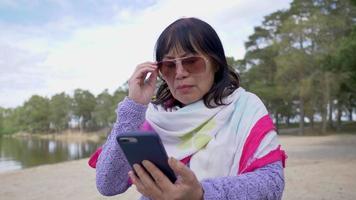 asiatische alte frau, die auf einem stuhl neben dem strand sitzt und ihr telefon benutzt, die sonnenbrille abnimmt und den bildschirm verschiebt, dann eine beschwerde einreicht und wütend wird video