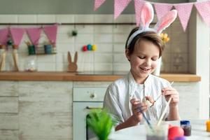 Close up of smiling boy sitting at table in kitchen and painting easter egg.