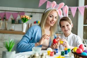 Funny, cheerful, joyful mum teaching, training her little son draw, paint, decorate easter eggs, together wearing bunny ears, preparing for Easter photo