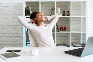 Mujer joven positiva sentada en una silla en una mesa con una computadora portátil poniendo sus manos detrás de su cabeza foto