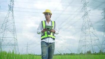 Font View.Electrical engineer wearing a Yellow helmet and safety carrying using tablet vest walking near high voltage electrical lines towards power station on the field.