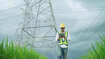 font view.electrical engineer che indossa un casco giallo e sicurezza che trasporta utilizzando giubbotto tablet camminando vicino a linee elettriche ad alta tensione verso la centrale elettrica sul campo. video