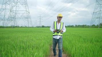 font view.electrical engineer che indossa un casco giallo e sicurezza che trasporta utilizzando giubbotto tablet camminando vicino a linee elettriche ad alta tensione verso la centrale elettrica sul campo. video