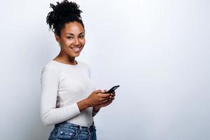Chica atractiva sobre un fondo de una pared blanca sonriendo con un teléfono en sus manos foto
