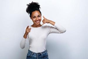 Positive curly girl in casual clothes shows thumb in camera and gesture call me looks into camera and smiles on white background