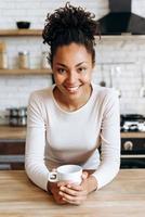 Hermosa mujer en ropa casual está sosteniendo una taza, mirando a la cámara y sonriendo mientras está sentada en la cocina foto