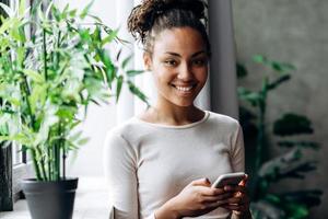 Hermosa chica afroamericana en ropa casual usa smartphone y sonríe mientras está de pie junto a la ventana foto