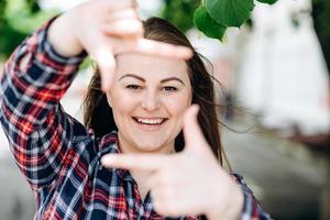 Portrait of playful beautiful caucasian woman pretending to take photo with her hands while walking on the street. Attractive model posing, using hands to make a frame outdoors