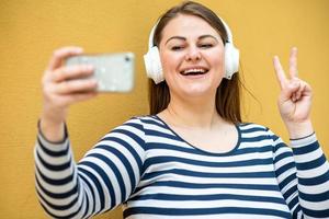 Against the background of an orange wall, a cheerful, smiling woman shows a gesture of peace and takes a selfie on a smartphone