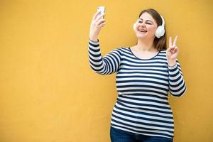 Against the background of an orange wall, a cheerful, smiling woman shows a gesture of peace and takes a selfie on a smartphone photo