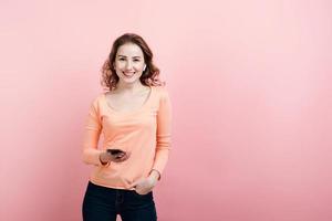 Retrato de mujer joven sonriente sosteniendo un teléfono móvil en la mano, usando audífonos inalámbricos, mirando a la cámara, vistiendo una camisa casual, aislado sobre fondo rosa foto