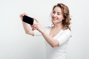 Smiling girl on white wall background makes selfie on phone photo