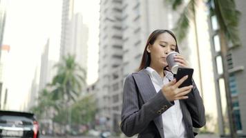 Empresaria asiática caminando en la calle mientras usa un teléfono inteligente y sostiene una taza de café video