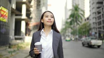 donna d'affari asiatica che cammina per strada mentre usa uno smartphone e tiene in mano una tazza di caffè