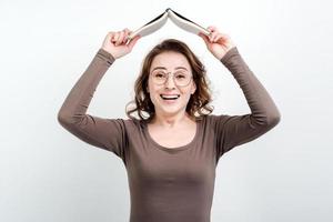 Happy funny woman in glasses  standing against a studio background holding a book above her head photo