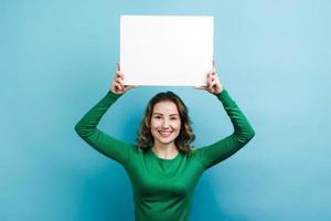 Chica rizada vistiendo un suéter verde sosteniendo un espacio de copia de pizarra blanca sobre su cabeza contra el fondo azul. foto