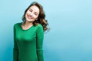 Cute blonde girl with curly hair and wearing in green sweater happily smiling against blue background photo
