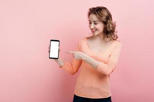 una niña sonriente sostiene un teléfono en la mano y señala con el dedo una pantalla blanca. aislado en un fondo rosa. foto