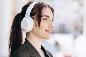 Foto de mujer agradable alegre usando auriculares tecnología moderna de pie en el fondo de la ciudad