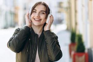Adorable lady in standing in city and listening favorite song in headphones. Outdoor portrait of dreamy european girl wears white earphones. photo