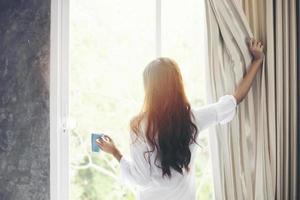 mujeres asiáticas tomando café y se despiertan en su cama completamente descansadas y abren las cortinas por la mañana para tomar aire fresco bajo el sol foto