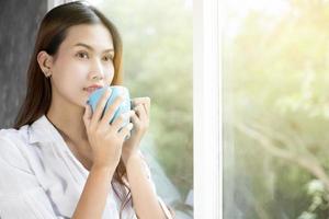 mujeres asiáticas tomando café y se despiertan en su cama completamente descansadas y abren las cortinas por la mañana para tomar aire fresco bajo el sol foto