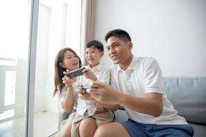familia asiática divirtiéndose jugando juegos de consola de computadora juntos, padre e hijo tienen los controladores del teléfono y la madre anima a los jugadores. foto