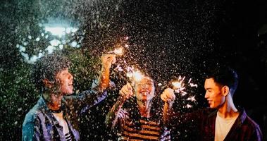 Grupo asiático de amigos con barbacoa en el jardín al aire libre riendo con bebidas alcohólicas de cerveza y mostrando un grupo de amigos divirtiéndose con bengalas en la noche, enfoque suave foto