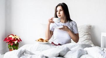 Joven mujer morena sentada despierta en la cama con globos en forma de corazón rojo y adornos bebiendo café comiendo croissants