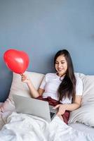 Feliz joven mujer morena sentada en la cama con globos en forma de corazón rojo trabajando en la computadora portátil saludando a amigos