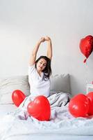 Feliz joven mujer morena sentada despierta en la cama con globos en forma de corazón rojo foto