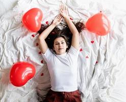 Joven mujer morena feliz acostado en la cama con globos y decoraciones en forma de corazón rojo